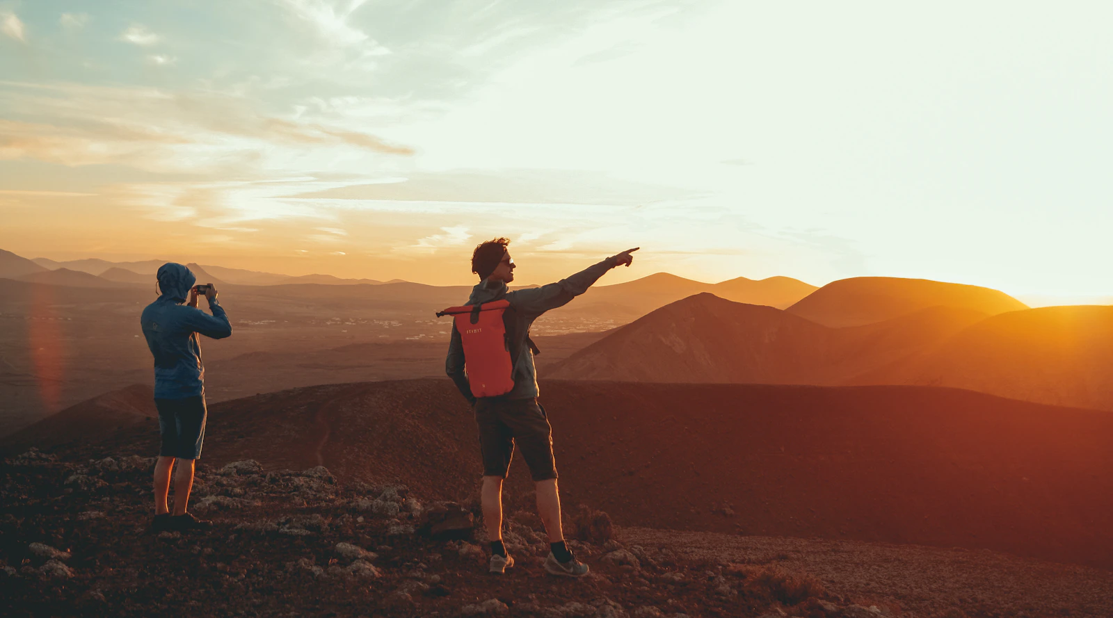 Two hikers on a mountain at sunset, one pointing towards the horizon